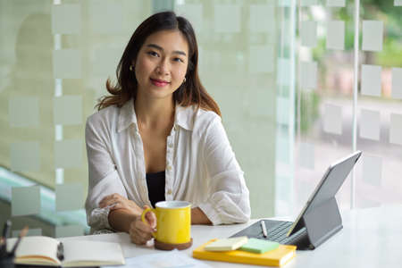 Attractive asian young businesswomen, female entrepreneur sits at her office work station with portable tablet and stuff on the tableの写真素材