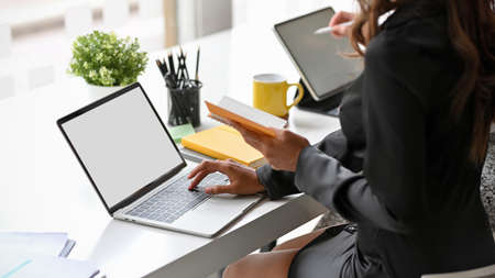 Cropped of elegant secretary working on laptop computer and checking some informations on her notebook at the desk.の写真素材