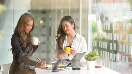 Two businesswomen working peacefully and relaxedly in an office co-working space while sipping morning coffee together.の写真素材