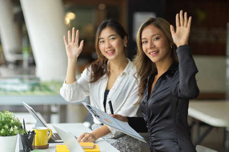 Beautiful and cheerful businesswomen raising hand palm up, hand greeting, saying hi or hello, hand gesture at her office space.の写真素材