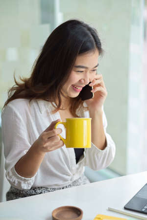 Portrait of cheerful businesswoman drink coffee while having a good talk on the phone at office.の写真素材