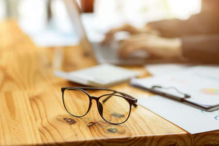 Close-up image of eye-glasses on wooden table with blurred businesspeople working at the office in the background.の写真素材