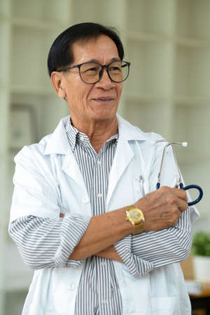 A successful Asian middle-aged male therapist with eyeglasses and a white gown stands with his arms crossed in his clinic.の写真素材