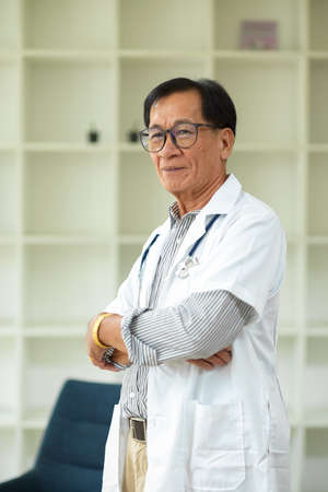 Portrait of a middle-aged male specialist and successful doctor in a white medical gown with his arms crossed in the office.の写真素材