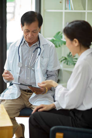 Middle-aged male specialist doctor in medical white gown examine diagnosis female patient at hospital office.の写真素材