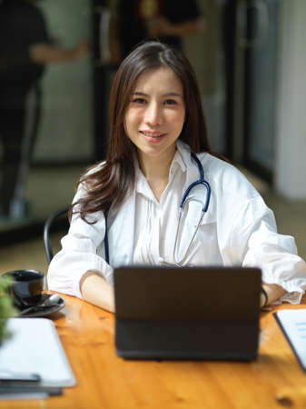 Portrait of a cheerful asian female doctor sits at her working desk in office, working on tablet touchscreen.の写真素材