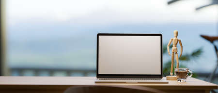 Laptop computer blank screen mockup on wooden table with coffee cup, wood figure and empty space for montage over blurred mountain view background. 3d rendering, 3d illustrationの写真素材