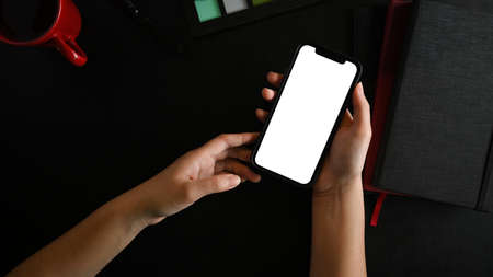 Close-up female hands holing mobile phone white screen mockup on black worktable. black background. top view, flat layの写真素材