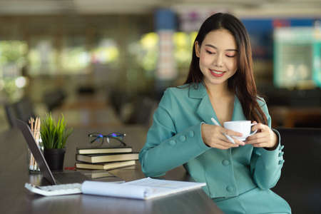 Attractive and confident businesswoman, female entrepreneur in stylish suit sitting in a office and looking at financial data while sipping coffee.の写真素材