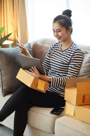 A woman online seller checks her inventory on her laptop computer before packing her product in a...
