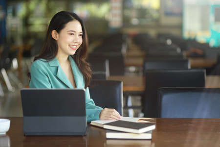 Beautiful millennial woman in stylish suit sitting with digital tablet computer and books in...