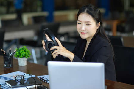 Beautiful young businesswoman holding headphone while looking at laptop computer screen, working at her office desk.の写真素材