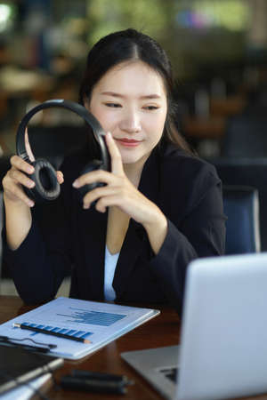 Beautiful asian businesswoman holding headphone while looking at laptop screen at her workplace.の写真素材