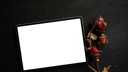 Tablet touchscreen mockup in white empty screen with dry roses on black background. modern dark worktable. top viewの写真素材