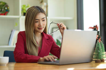 Elegant asian young business woman in red suit holding eyeglasses while working on laptop computer at office desk.の写真素材