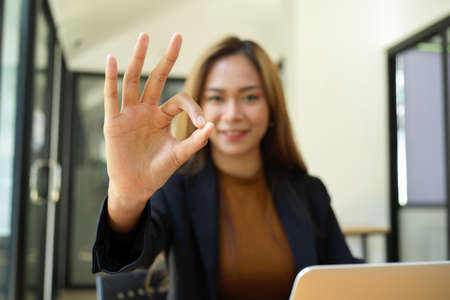 Cheerful asian young business worker posting OK hand sign at her desk. deal, agreement, happy, ok gesture.の写真素材