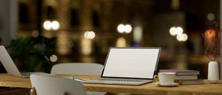 A wooden desk with notebook laptop computer white screen mockup over blurred the street in the evening on background. 3d rendering, 3d illustrationの写真素材