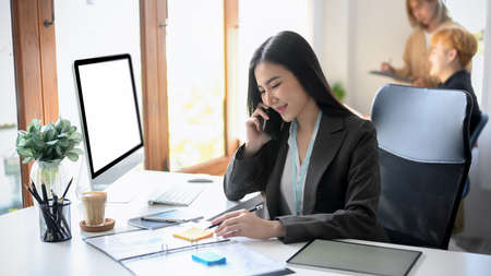 A beautiful asian businesswoman or female secretary talking on the phone with business partners at her modern office desk.の写真素材
