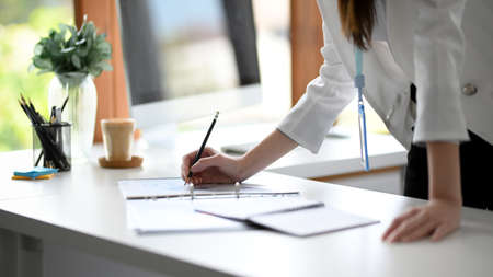 Cropped image of a professional businesswoman working on a financial report and writing on paperwork while leaning on a desk.の写真素材