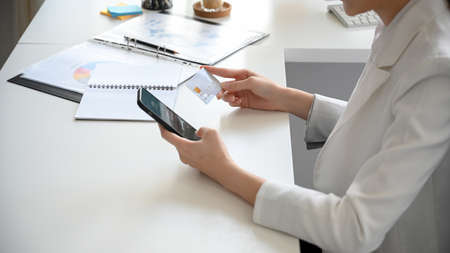 Side view of a female employee or businesswoman holding smartphone and credit card at her desk. Mobile banking, online payment concept.の写真素材