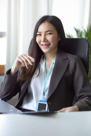 Portrait image of a beautiful, cheerful and friendly Asian business woman in a formal suit sitting at her desk in a office workplace.の写真素材