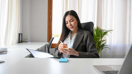 an Asian young business woman reading a document and taking a memo notes on a sticky notes at her office desk. business woman working at her table.の写真素材