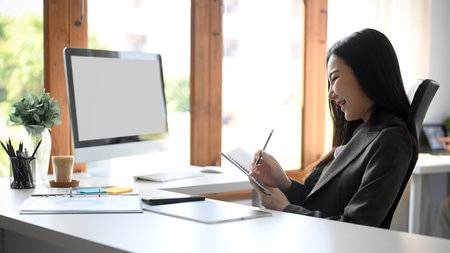 Cheerful asian young business woman sitting at her desk and writing something on her personal notebook.の写真素材