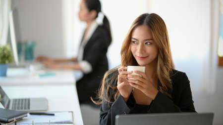An attractive female marketing employee sipping a morning coffee and looking out the window at her desk.の写真素材