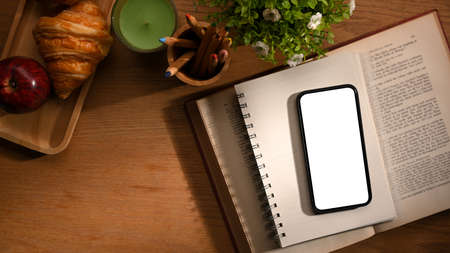 Cozy vintage worktable with smartphone white display mockup on a book, stationery, decor, a breakfast set croissant and apple on wooden table. top viewの写真素材