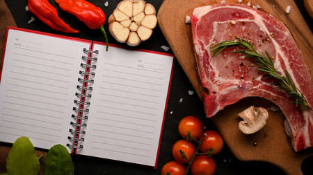 Raw pork chop steak with special seasoning on wooden board and an open notebook with blank pages for...