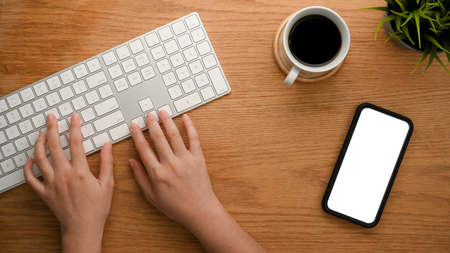 Close-up, top view of a girl's hands typing on desktop computer keyboard, smartphone white screen on wooden table.の写真素材