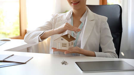 Cropped shot of a beautiful property realtor or real estate agent holding a house model at her office desk.の写真素材