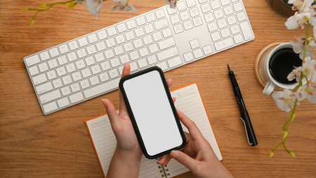 Top view of a smartphone white screen for display your graphic ads on a woman's hands over workspaceの写真素材