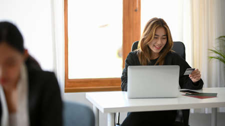 Happy Asian young businesswoman with a credit card, sitting at her office desk. Online shopping,...