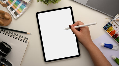 Graphic designer workspace concept with female hands using stylus pen sketching on digital tablet computer. watercolour palette, colour swatch samples, camera and stationery on white table.の写真素材