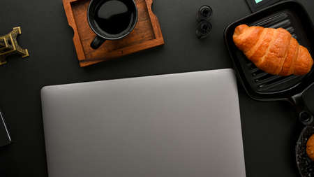 Minimal dark office desk with laptop computer, coffee and croissant on black background. top view