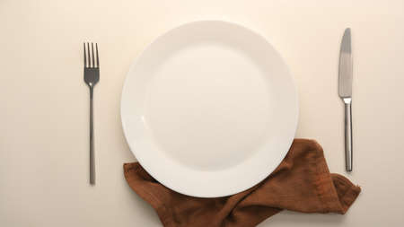 Top view of a blank white plate with silverware and brown napkin over white dining table. empty plate mockup.の写真素材