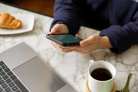 Close-up, A woman in sweater sitting at her worktable and using modern smartphone in the morning. A young female spending time on online shopping website.の写真素材
