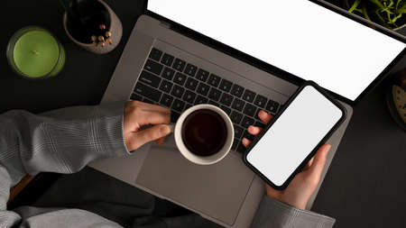 Top view of a businesswoman using her smartphone while sipping morning coffee at her office desk.
