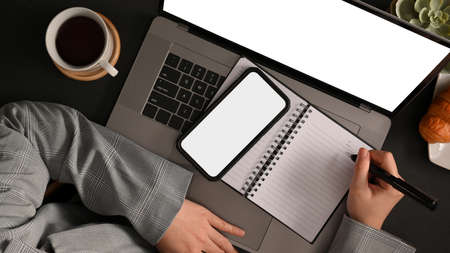 Top view of a busy businesswoman working on laptop computer, looking some details on smartphone...