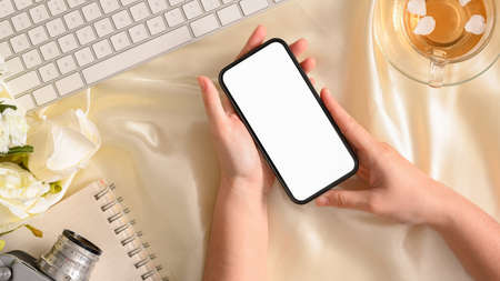 Beautiful stylish feminine workspace with female's hands holding a smartphone white screen mockup. top viewの写真素材