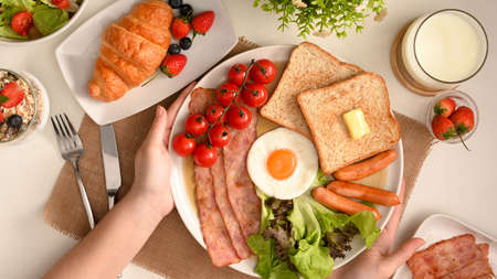 Top view, flat lay, Close-up a female's hands serving a breakfast set on dining table. egg, ham, toasts, sausages, croissant, salad and milkの写真素材