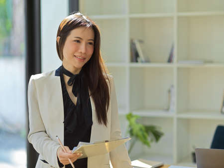 Portrait of Asian gorgeous businesswoman in formal suit, standing in the office and holding a clipboard.の写真素材
