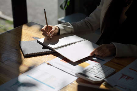 Cropped shot of a female manager or businesswoman taking notes on her personal notebook and working on financial documents in the bright office.の写真素材