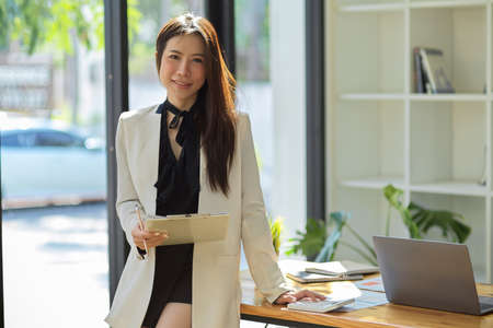 Confident and successful businesswoman in stylish outfit with clipboard paper, leaning on table and smiling to camera in the modern office.の写真素材