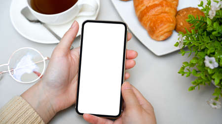 A young female hand holds a smartphone white screen mockup over a coffee table with a coffee cup and...