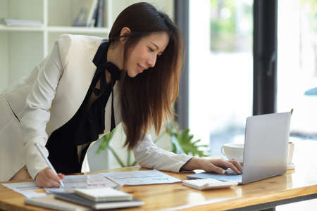 Asian female boss checking her financial project proposal on laptop computer at her desk in the office. Businesswoman leaning on table and looking at laptop screen.の写真素材