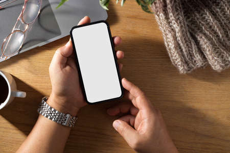 A male's hand holding a smartphone white screen mockup over wooden worktable. top view, flat layの写真素材