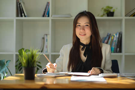 Beautiful Asian young businesswoman taking notes in her modern office workplace, checking report informations, reading paperworks.の写真素材