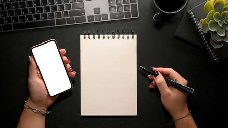 A female looking some informations on mobile phone screen and taking list on spiral notepad on modern dark office desk. closeup, top viewの写真素材